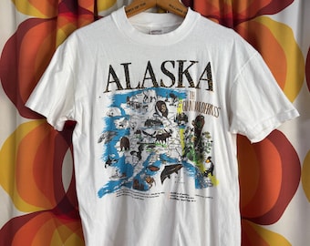 May include: A white short-sleeve t-shirt featuring a graphic map of Alaska with the text ALASKA THE GIANT WILDERNESS. The design includes illustrations of various wildlife, including a bear, whale, and eagle, along with printed facts about the state.