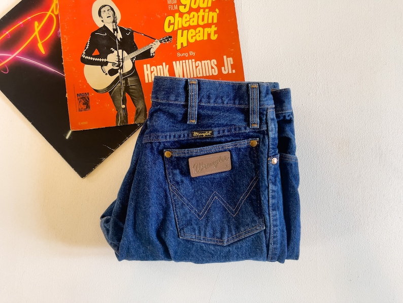Puede incluir: Un par de pantalones vaqueros azules de mezclilla con un parche de cuero marr&oacute;n que dice "Wrangler" en el bolsillo trasero.