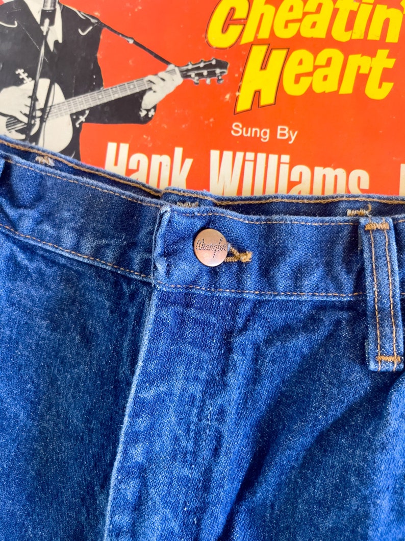 Puede incluir: Primer plano de un par de pantalones vaqueros azules de mezclilla con un bot&oacute;n Wrangler en la cintura.