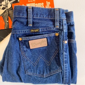 Puede incluir: Un par de pantalones vaqueros de mezclilla azul oscuro con un parche de cuero marr&oacute;n Wrangler en el bolsillo trasero.