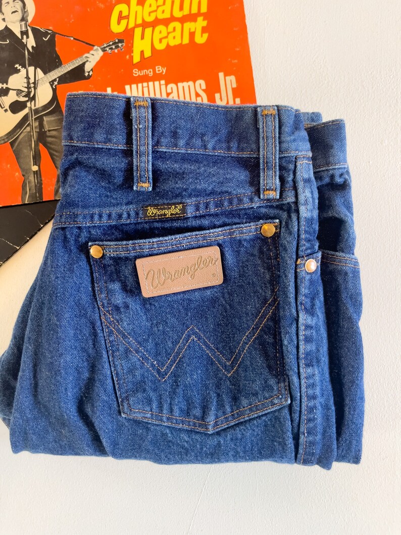 Puede incluir: Un par de pantalones vaqueros de mezclilla azul oscuro con un parche de cuero marr&oacute;n Wrangler en el bolsillo trasero.
