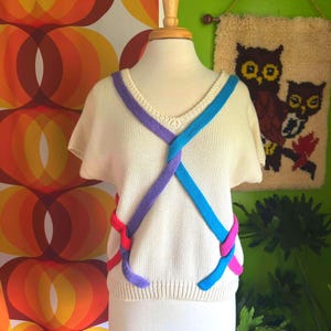 May include: A cream-colored knitted sweater with short sleeves and a V-neck. The sweater has a unique design with colorful braided straps in pink, blue, and purple.