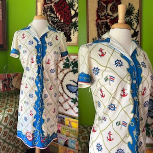 May include: A vintage nautical-themed dress on a mannequin. The dress features a white background with a repeating pattern of red anchors, blue ship wheels, and flags. A blue stripe runs down the front, and the dress has short sleeves.