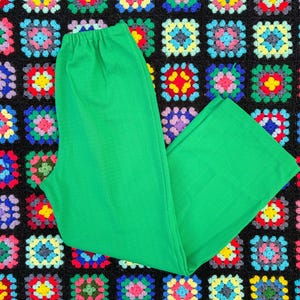May include: Bright green pants with an elastic waistband, lying on a colorful, patterned crochet blanket. The pants are a solid, vibrant green color. The blanket features a repeating pattern of square crochet blocks in various colors.