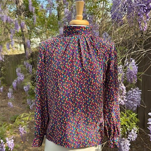 May include: A vintage, long-sleeved blouse with a high neck and a colorful, abstract print. The print features small, multi-colored dots and shapes on a dark blue background.