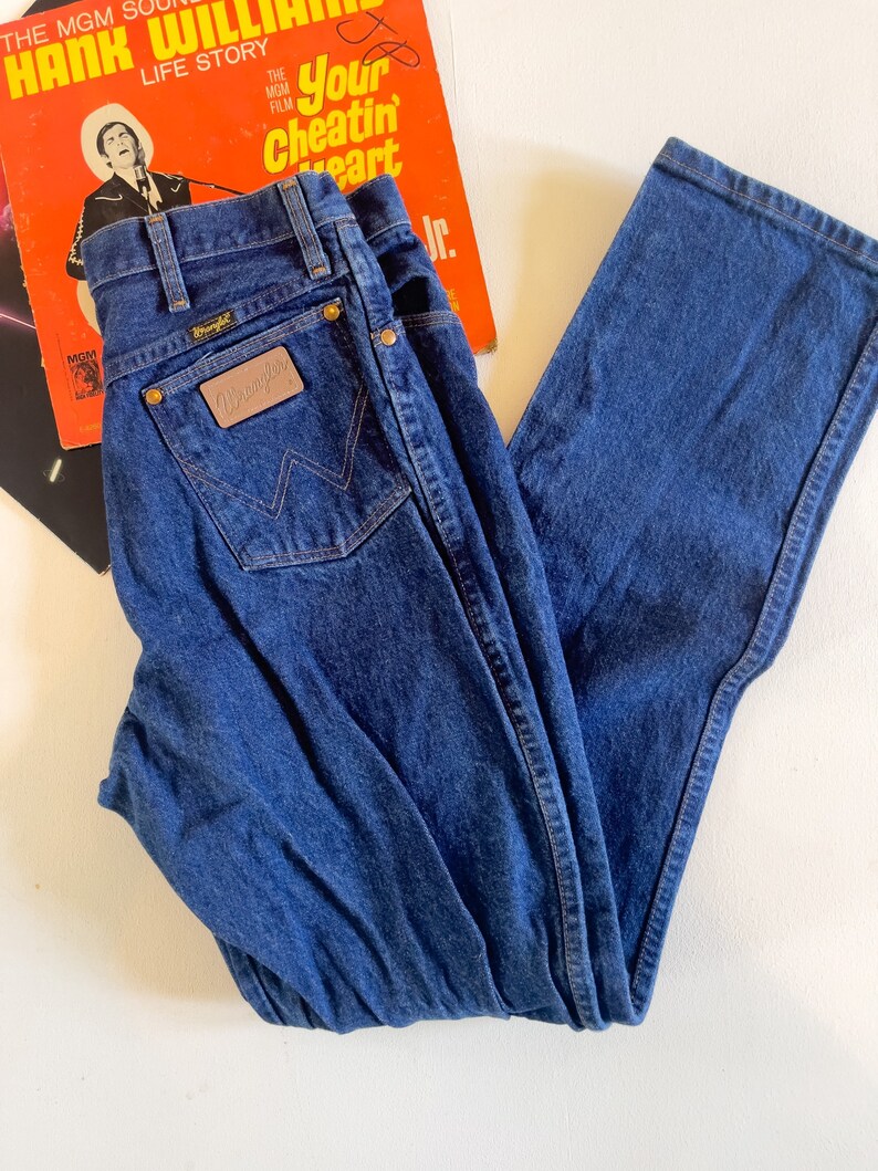 Puede incluir: Un par de pantalones vaqueros de mezclilla azul oscuro con un parche de cuero marr&oacute;n que dice "Wrangler" en el bolsillo trasero.