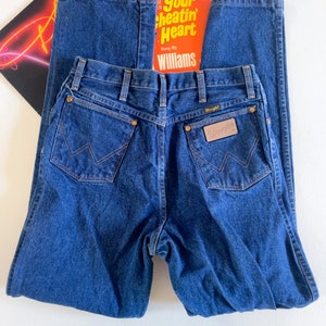 Puede incluir: Un par de pantalones vaqueros de mezclilla azul oscuro con una etiqueta Wrangler de cuero marr&oacute;n en el bolsillo trasero. Los pantalones tienen un estilo cl&aacute;sico de pierna recta.