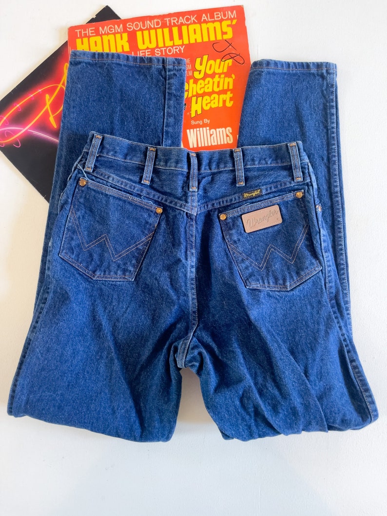 Puede incluir: Un par de pantalones vaqueros de mezclilla azul oscuro con una etiqueta Wrangler de cuero marr&oacute;n en el bolsillo trasero. Los pantalones tienen un estilo cl&aacute;sico de pierna recta.