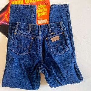 Puede incluir: Un par de pantalones vaqueros de mezclilla azul oscuro con una etiqueta Wrangler de cuero marr&oacute;n en el bolsillo trasero.