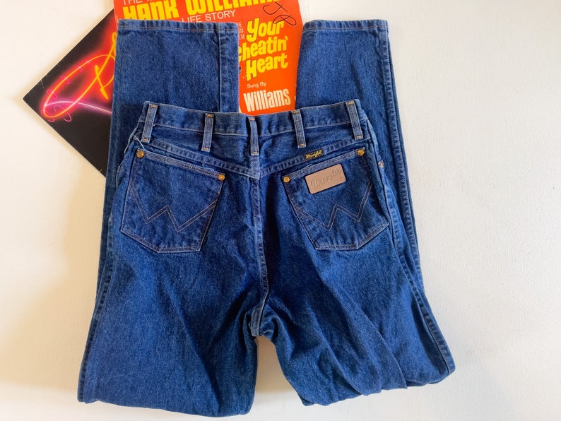 Puede incluir: Un par de pantalones vaqueros de mezclilla azul oscuro con una etiqueta Wrangler de cuero marr&oacute;n en el bolsillo trasero.