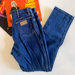 Puede incluir: Un par de pantalones vaqueros azules de mezclilla con un parche de cuero marr&oacute;n que dice "Wrangler" en el bolsillo trasero.
