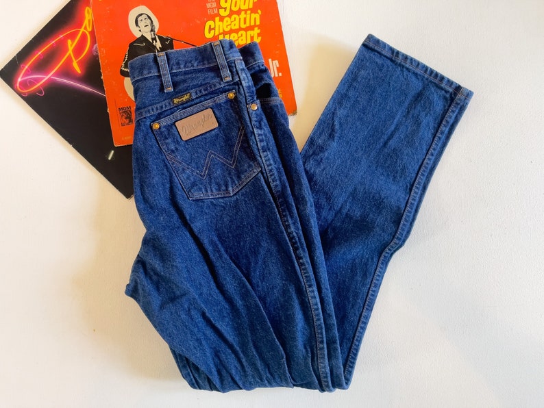 Puede incluir: Un par de pantalones vaqueros azules de mezclilla con un parche de cuero marr&oacute;n que dice "Wrangler" en el bolsillo trasero.