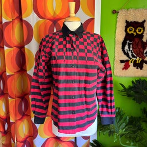May include: A red and black checkered button-up shirt with long sleeves. The shirt is on a mannequin against a colorful background.
