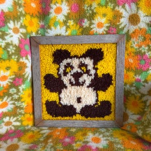 May include: A square wall hanging featuring a textured punch needle or latch hook panda bear design in dark brown and cream yarn against a bright yellow background, enclosed within a simple, weathered light brown wooden frame.