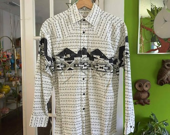 May include: A long-sleeved button-down shirt with a white base and a repeating black and gray pattern. The shirt features a geometric design across the chest and shoulders, with black buttons and cuffs. The shirt is hanging on a wooden hanger.