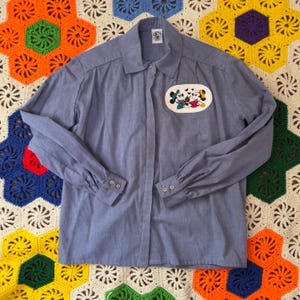 May include: A light blue button-down shirt with long sleeves. The shirt features an embroidered patch of Mickey and Minnie Mouse. The shirt has a collar and buttoned cuffs. The shirt is made of a lightweight fabric.