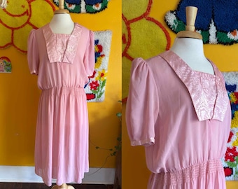 May include: A vintage pink dress with short sleeves and a square neckline. The dress features a textured, patterned collar and a gathered waist. The skirt falls to the mid-calf. The dress is displayed on a mannequin against a yellow background.