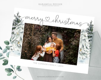 May include: A Christmas card featuring a family portrait with a white background and a watercolor illustration of snowy trees on either side. The text "merry christmas" is written in cursive with a heart symbol. The family is standing in front of a green bush. The text "THE CONNOR FAMILY | 20YY" is at the bottom of the card.