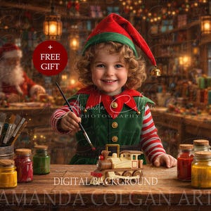 May include: A child dressed as an elf in a green and red outfit with a matching hat, holding a paintbrush over a small wooden toy train. The scene includes glass jars of colorful paint, paintbrushes in a metal container, and a red circle with the text + FREE GIFT.