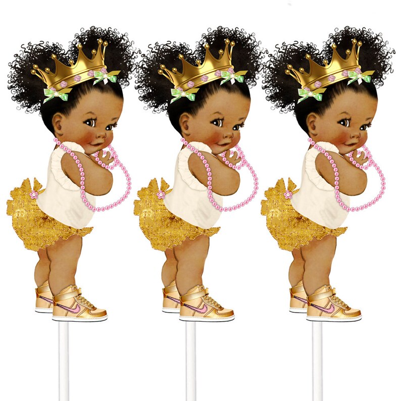 May include: Three cupcake toppers featuring a cartoon illustration of a Black baby girl wearing a gold crown, a white dress, a gold skirt, pink pearl necklace, and gold sneakers.