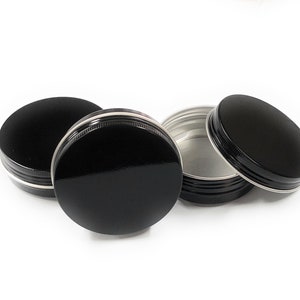 May include: Three black metal tins with silver rims. The tins are round and have a smooth, shiny finish. Two of the tins are closed, and one is open, revealing a silver interior.