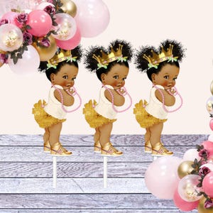 May include: Three gold and white cupcake toppers featuring a cartoon baby girl wearing a gold crown, a white dress, a gold tutu, and pink sneakers. The baby girls are holding pink pearl necklaces.