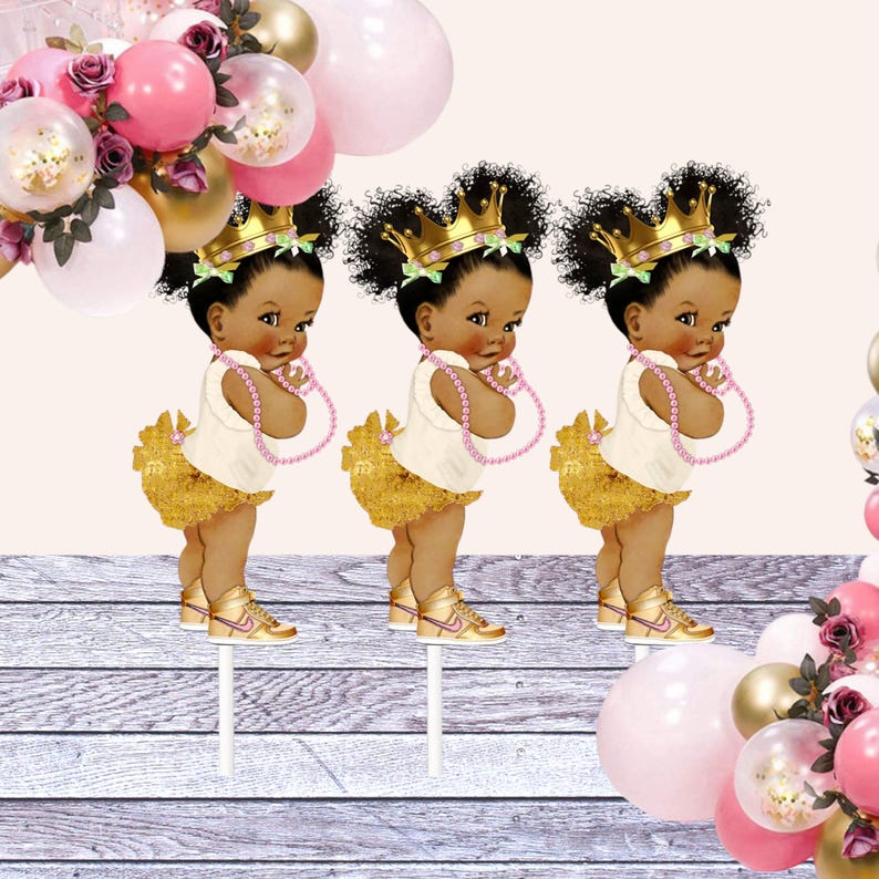 May include: Three gold and white cupcake toppers featuring a cartoon baby girl wearing a gold crown, a white dress, a gold tutu, and pink sneakers. The baby girls are holding pink pearl necklaces.
