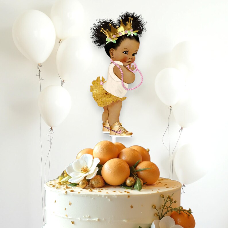 May include: A cake topper featuring a cartoon illustration of a little girl with brown skin, wearing a gold crown, a white shirt, a gold skirt, and pink shoes. The girl is holding a pink beaded necklace. The cake topper is on a white cake decorated with gold sprinkles and orange fruit.