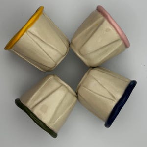 May include: Four small, off-white ceramic cups with textured sides and colourful rims. The rims are painted in yellow, pink, dark blue, and green. The cups are arranged in a circular pattern.