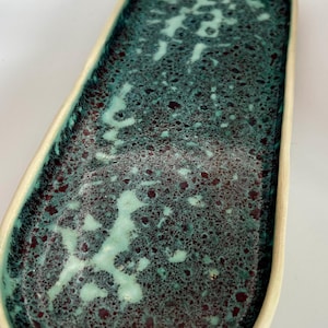 May include: An oval ceramic tray with a speckled teal and burgundy glaze. The tray has a cream-coloured rim and a textured surface. This decorative tray is suitable for serving or display.