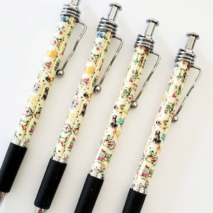 May include: Four ballpoint pens with a cream-colored barrel featuring a repeating pattern of dogs, flowers, and other small illustrations. The pens have silver-colored metal accents, black rubber grips, and are arranged diagonally on a white surface.