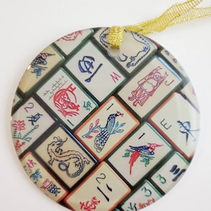 May include: Round glass ornament with a colorful design of Mahjong tiles. The tiles feature various symbols and images, including dragons, birds, and flowers. The ornament is decorated with gold ribbon.