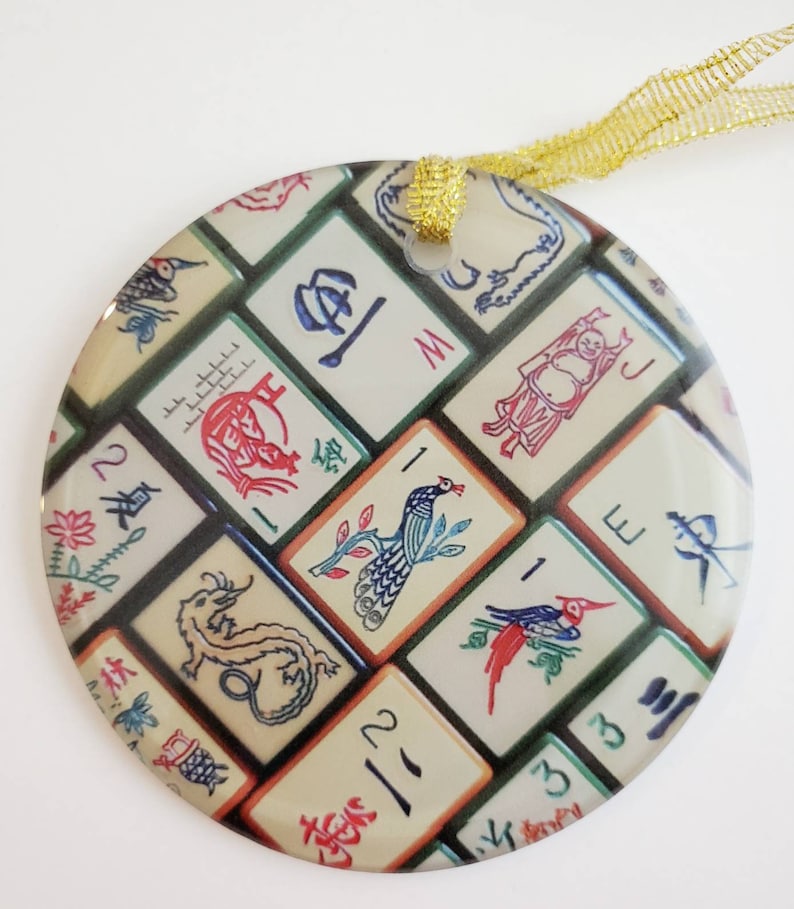 May include: Round glass ornament with a colorful design of Mahjong tiles. The tiles feature various symbols and images, including dragons, birds, and flowers. The ornament is decorated with gold ribbon.