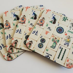 Op de afbeelding: Een set van vier vierkante onderzetters met een Mahjong-tegelontwerp. Elke onderzetter toont verschillende tegels met kleurrijke illustraties, Chinese karakters en cijfers. De onderzetters hebben een crèmekleurige achtergrond met een donkerbruine rand.