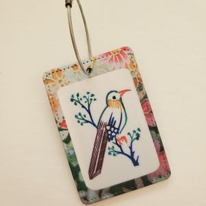May include: A rectangular luggage tag with a bird illustration. The tag features a white center with a colorful bird perched on a branch. The border has a floral pattern with pink, orange, and green hues. A silver-tone metal loop is attached.