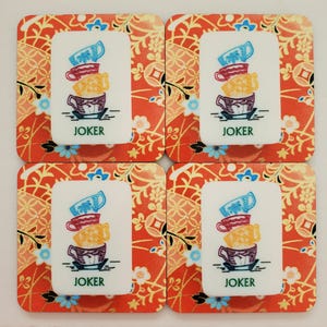 Puede incluir: Juego de cuatro posavasos cuadrados. Cada posavasos tiene un fondo floral rojo y dorado. Un rectángulo blanco en el centro de cada posavasos presenta una pila de tazas de té coloridas y la palabra "JOKER".