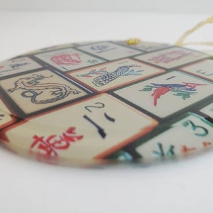 May include: Round glass ornament with a colorful image of Chinese mahjong tiles. The tiles feature various symbols and numbers, including a dragon, a bird, and the number 2.