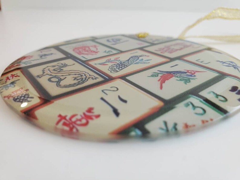 May include: Round glass ornament with a colorful image of Chinese mahjong tiles. The tiles feature various symbols and numbers, including a dragon, a bird, and the number 2.