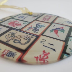 May include: Round glass ornament with a design of Chinese mahjong tiles. The tiles feature various symbols, numbers, and images, including a dragon, a peacock, and a bird. The ornament has a gold string for hanging.
