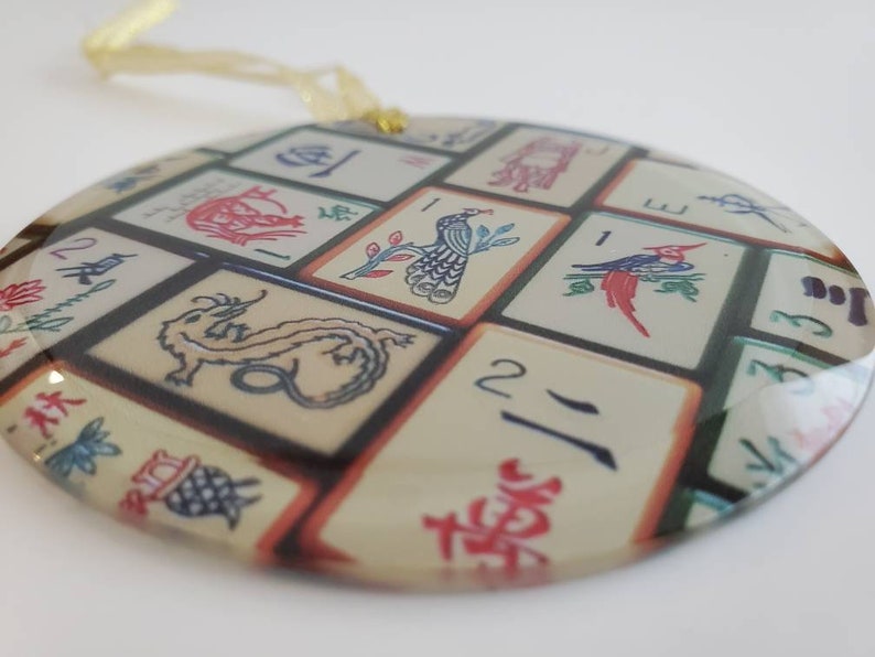 May include: Round glass ornament with a design of Chinese mahjong tiles. The tiles feature various symbols, numbers, and images, including a dragon, a peacock, and a bird. The ornament has a gold string for hanging.