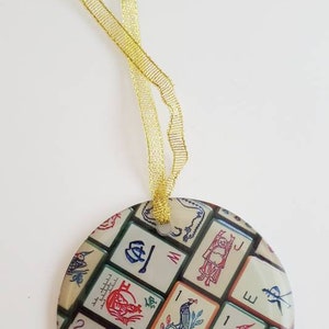 May include: Round, white, and green ornament with a gold ribbon. The ornament is decorated with a pattern of colorful Chinese mahjong tiles.