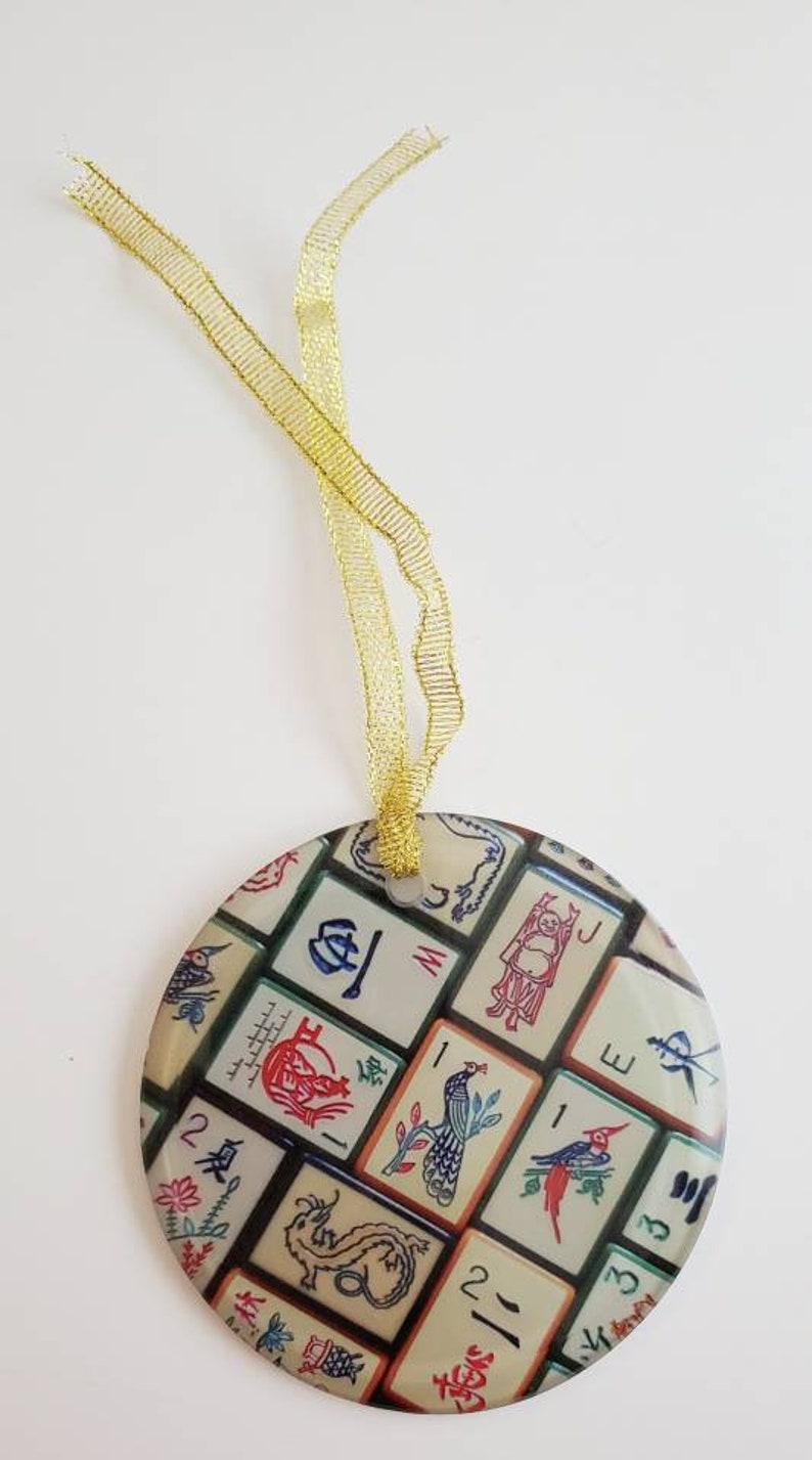 May include: Round, white, and green ornament with a gold ribbon. The ornament is decorated with a pattern of colorful Chinese mahjong tiles.