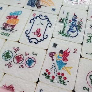 Puede incluir: Primer plano de un juego de fichas de Mahjong con varios dise&ntilde;os coloridos, incluyendo un p&aacute;jaro, un hada, mariposas y una pila de tazas. Las fichas son de cer&aacute;mica blanca con contornos negros y est&aacute;n dispuestas en una cuadr&iacute;cula.