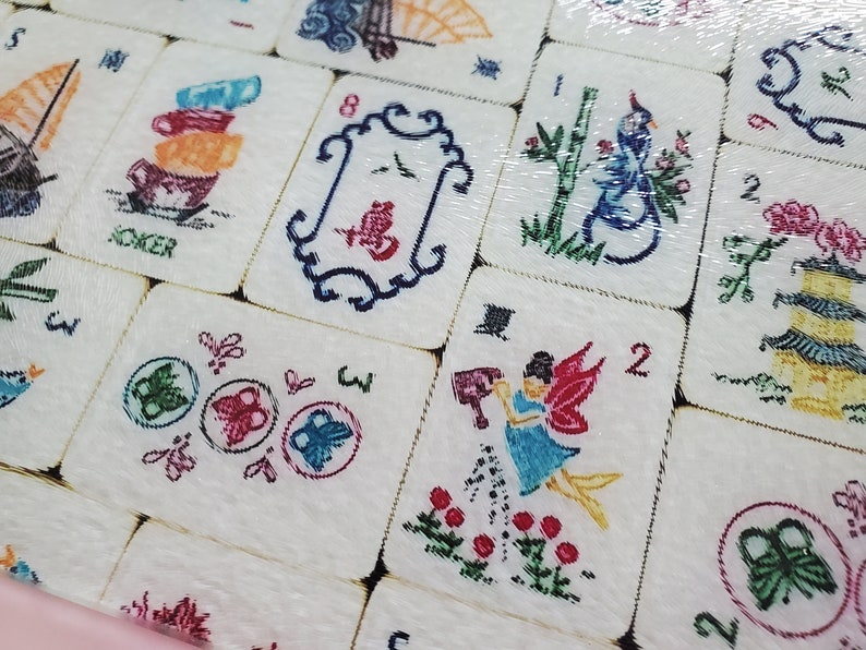 Puede incluir: Primer plano de un juego de fichas de Mahjong con varios dise&ntilde;os coloridos, incluyendo un p&aacute;jaro, un hada, mariposas y una pila de tazas. Las fichas son de cer&aacute;mica blanca con contornos negros y est&aacute;n dispuestas en una cuadr&iacute;cula.