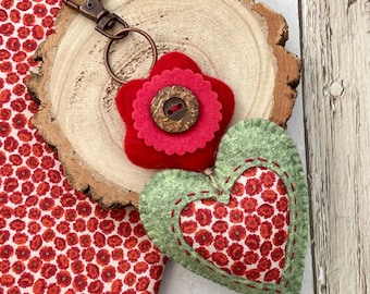 May include: A red felt flower with a brown button center attached to a green felt heart with a red floral pattern inside. The heart is attached to a key ring.