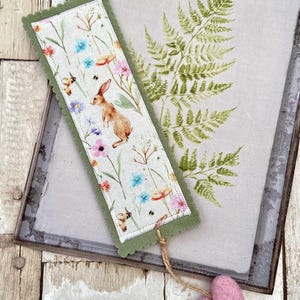 May include: A rectangular fabric bookmark featuring a printed pattern of a brown rabbit, colourful wildflowers, and small insects. The bookmark has a scalloped olive green felt border and a jute twine string attached to a small pink felt heart charm.