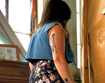 May include: A woman wearing a denim vest and a floral skirt stands in a room with a wooden wall and a dresser. She is wearing black boots and has a tattoo on her arm.
