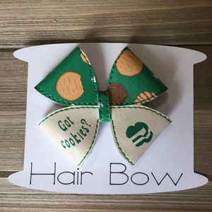 Könnte beinhalten: Grünes und braunes Haarband mit einer weißen Schleife mit einem Girl Scout-Logo und dem Text "Got Cookies?" darauf. Die Schleife befindet sich auf einer weißen Karte mit dem Text "Hair Bow" darauf.