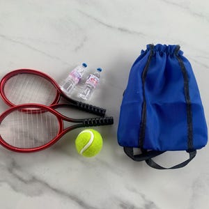 以下が含まれることがあります： 2つの赤いラケット、緑のテニスボール、2つの小さな水筒、青い巾着バックパックを備えたミニチュアテニスセット。ラケットは黒いハンドルと白いストリングスです。水筒は透明で青いキャップが付いています。