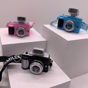 May include: Three miniature toy cameras in pink, blue, and black with silver accents and flash attachments. Each camera has a lens and a strap. The black camera has a strap that says "SMILE". The cameras are displayed on white blocks.